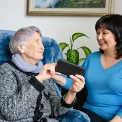 Illustrasjon: Eldre dame får opplæring i bruk av mobiltelefon av en yngre dame