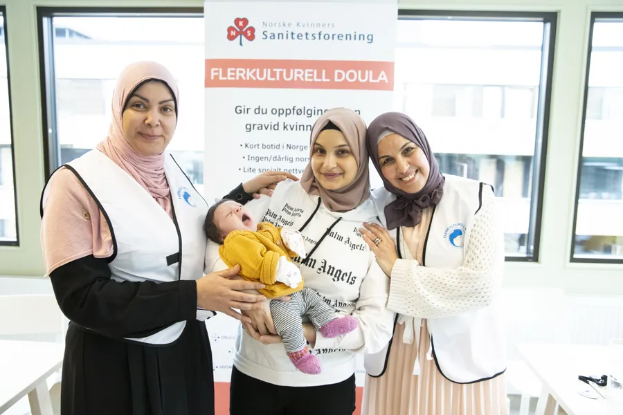 tre kvinner med hodeplagg. Kvinnen i midten holder en baby. De står foran et skilt som forklarer om flerkulturell doula.De smiler til kameraet