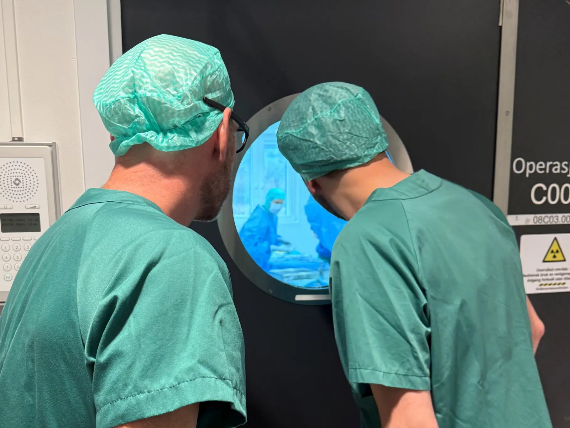 Et par menn iført kirurgiske uniformer som ser inn gjennom vindu til operasjonssal