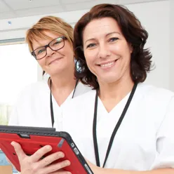 Bilde av to sykepleiere på et pasientrom. Smiler til kamera.